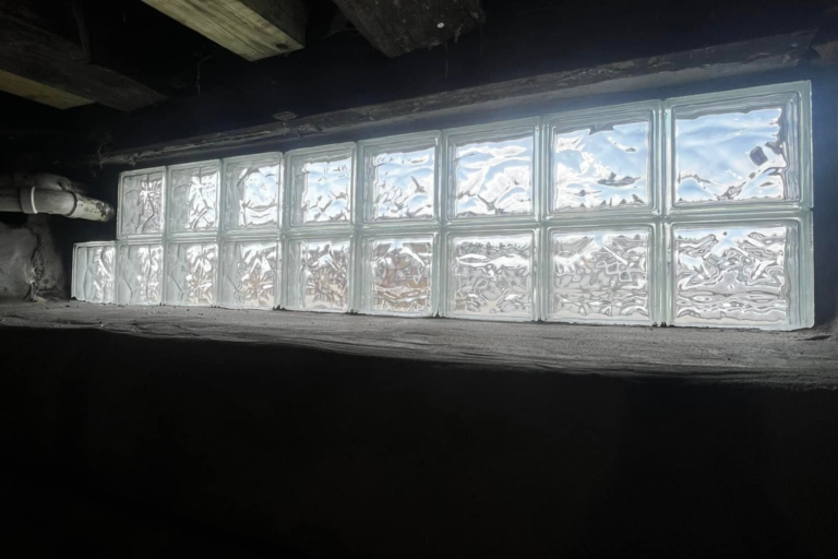 A window featuring a glass block, providing light and privacy in a crawl space setting.